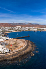 Fototapeta premium Aerial view of a crescent bay in Lanzarote, Canary Islands, with whitewashed resorts around a marina, a breakwater, a small lighthouse marker, and rust red calderas.