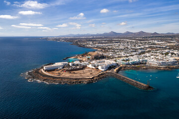 Aerial view of Costa Teguise, Lanzarote, shows curved breakwater, rocky shore, turquoise coves, hotel complexes, and a sandy construction area under clear daylight skies. © Aerial Film Studio