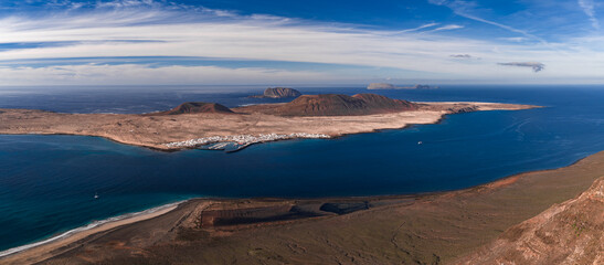 Aerial panorama shows La Graciosa from Mirador del Rio, Caleta de Sebo, rust volcanic cones, Chinijo islets, sailboat and ferry wake, rugged Lanzarote coast in daylight.