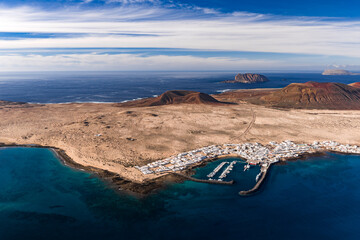 Aerial view of Caleta de Sebo, La Graciosa, shows whitewashed village, small marina, ochre sands, cobalt Atlantic, lava hills, and Isla de Montana Clara and Alegranza. © Aerial Film Studio
