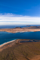 Aerial La Graciosa off Lanzarote shows Caleta de Sebo harbor, whitewashed houses, sandy beach, lone sailboat, lava fields, Montana Amarilla, islets, and Atlantic under streaked clouds. © Aerial Film Studio