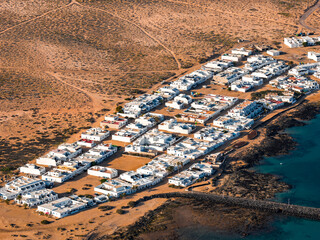 Aerial view of Caleta de Sebo on La Graciosa shows white flat roof homes, sandy tracks, and dark lava rock by turquoise Atlantic waters in warm late afternoon light. © Aerial Film Studio