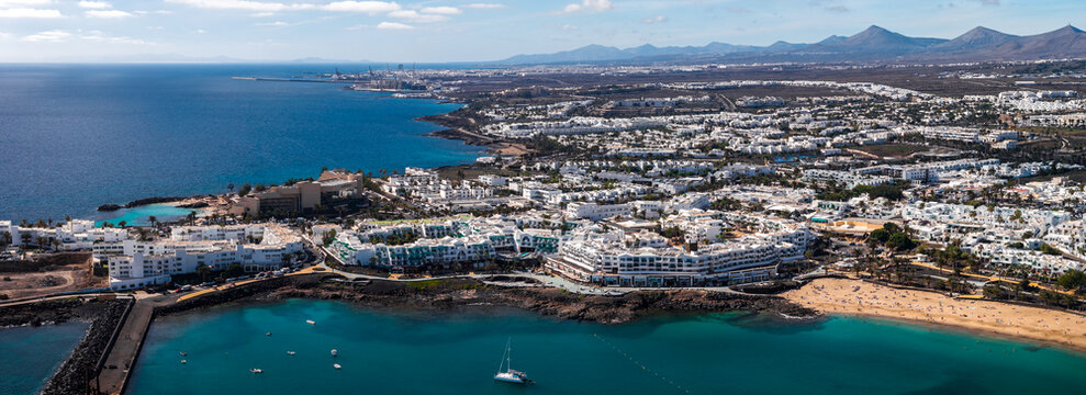 Aerial view shows Costa Teguise, Lanzarote, whitewashed buildings, sandy beaches, and boats. Frame looks north to Arrecife port and cranes with volcanic ridge beyond.