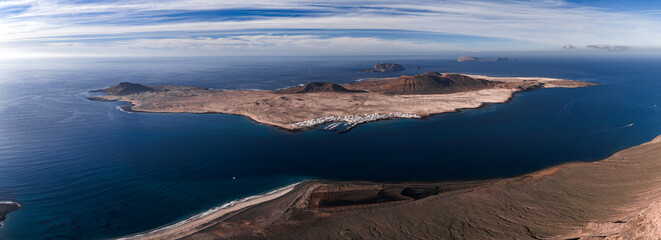 Aerial view shows La Graciosa with Caleta de Sebo harbor, volcanic cones, lava fields, Mirador del Rio on Lanzarote, cirrus bands, and calm turquoise shallows at midday.
