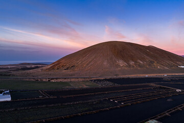 Naklejka premium La Geria vineyards with black lava plots and stone walls sit below a volcanic cone in Lanzarote, Canary Islands. A road with cars skirts the base, Atlantic on horizon.