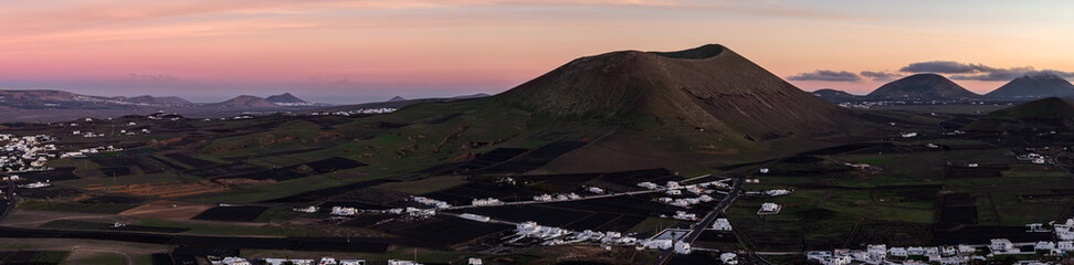 Aerial dusk view shows Lanzarote cinder cone above black lapilli fields. Whitewashed villages sit in La Geria with stone windbreaks, facing Timanfaya cones. © Aerial Film Studio