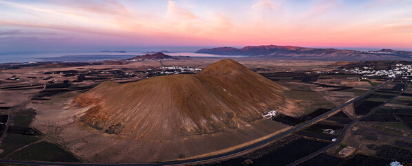 Aerial view at sunset of a volcanic cone in Lanzarote, La Geria terraces, black lava fields, and white villages, facing the Famara massif and La Graciosa near Mirador del Rio. © Aerial Film Studio
