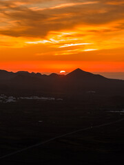 Fiery orange and gold clouds streak the sky as the sun sets over Lanzarote. A conical volcanic peak and Timanfaya calderas form silhouettes with whitewashed villages. © Aerial Film Studio