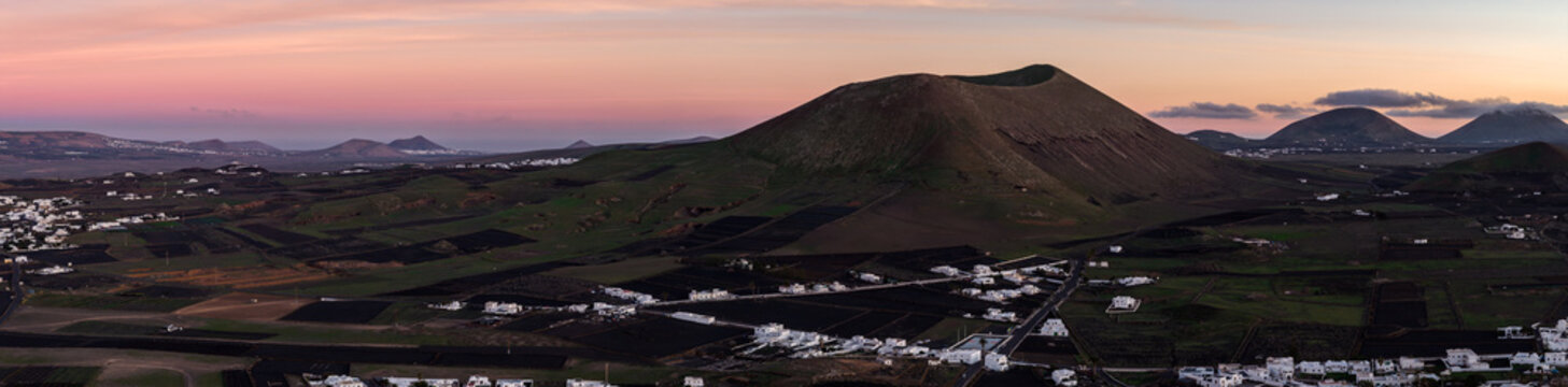 Aerial dusk view shows Lanzarote cinder cone above black lapilli fields. Whitewashed villages sit in La Geria with stone windbreaks, facing Timanfaya cones.