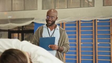 Waist up shot of smiling Black man as coach or teacher tracking time and taking notes on clipboard in PE class with children doing fitness exercises in foreground