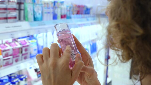 Close up of a young woman's hands holding a bottle of pink micellar water or toner, carefully reading the ingredient list and product information on the label in a beauty store aisle