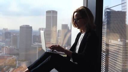 Silhouette of a successful businesswoman with glasses sitting on a windowsill in a modern office, typing on her laptop computer with a stunning view of the cityscape in the background - Powered by Adobe