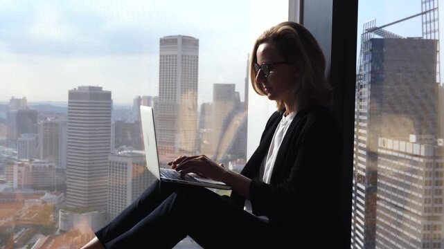 Silhouette of a successful businesswoman with glasses sitting on a windowsill in a modern office, typing on her laptop computer with a stunning view of the cityscape in the background
