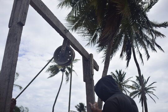 Homens trabalhando em po&ccedil;o, puxando corda em roldana de ferro em ambiente praiano com coqueiro de fundo