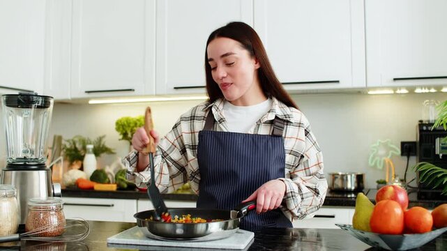 Young woman samples vegetables from frying pan while cooking in home kitchen for diet plan and daily wellness. Girl looks satisfied after taste test ready to serve homemade lunch in apartment indoors.