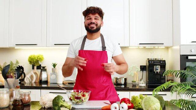 Arabian man jokes with pepper rings on eyes while mixing salad in kitchen for keto diet and clean eating. Young guy laughs feeling playful and proud after healthy home meal prep in apartment indoors.