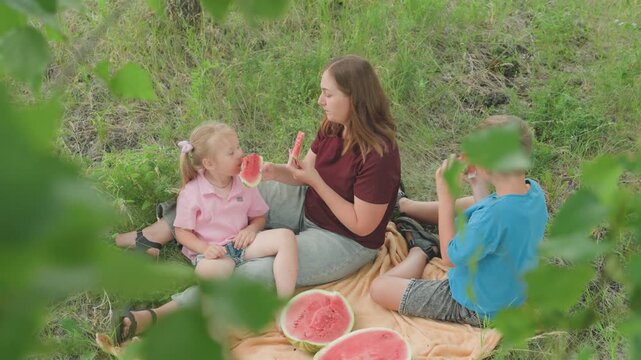 parent kids savor watermelon in meadow, picnic blanket with sliced melon and basket, leafy foreground framing candid smiles, siblings sharing slices and playful banter, summer warmth and relaxed