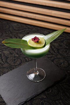 Enjoy a green cocktail with lime and leaves served on a black slate plate in a modern setting