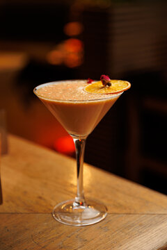 Cocktail served in a clear glass with dried orange garnish on a wooden table in a dimly lit bar setting