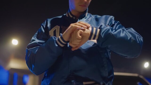 neon street man adjusting watch, ready for night shift club promoter and courier energy with quick ritual of wrist checks and jacket zips, folding hands and tapping time, blue varsity jacket