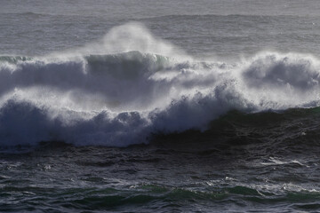 Naklejka premium A dark blue colored rip curl of a wave crashing on shore. The dangerous ocean swell has white foam and a water spray. There's a break in the center of the stormy surf with foam on both sides.