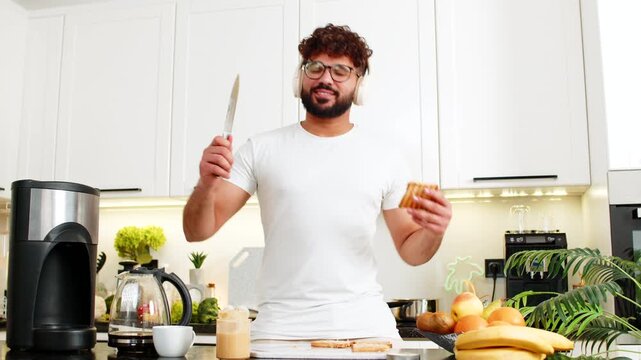 Arabian man listens to music and dances while spreading peanut butter on toast in home kitchen. Young guy enjoys joyful morning breakfast and coffee aroma after workout for healthy nutrition energy.