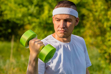 A man in a white t-shirt and headband lifts a green dumbbell outside on a sunny day.