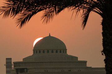 Obraz premium The sun sets behind a dome structure while a palm tree stands in the foreground. The scene captures the end of the day with warm colors in the sky.