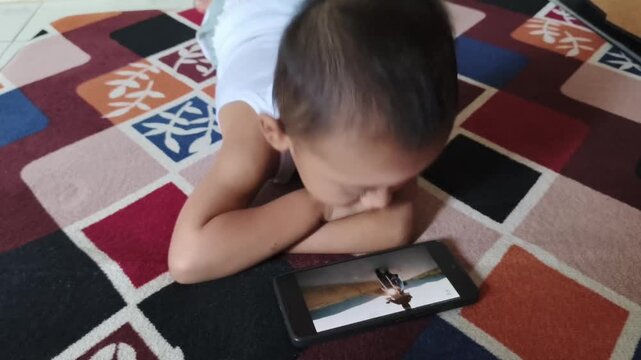 Young Asian boy lying prone on carpet watching video on smartphone wearing white sleeveless undershirt at home for digital generation and child technology use concept