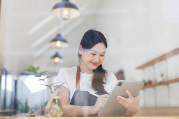 Portrait of happy asian woman using digital tablet in her store. 
Successful sme small business owner in casual wearing blue apron at entrance cafe.