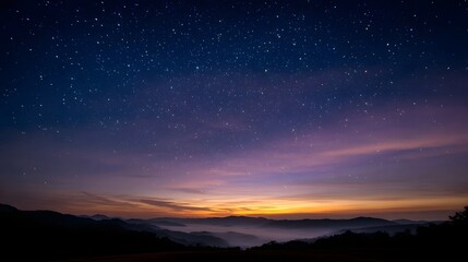 Naklejka premium Vast starry sky illuminates a misty mountain landscape at twilight with vibrant colors on the horizon