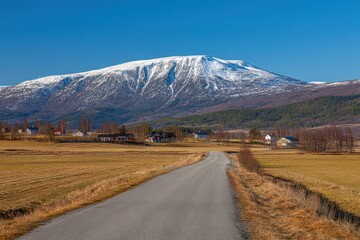 Picturesque Snow Mountain Under Clear Blue Sky with Winding Road Leading to Small Village with Red Houses in Rural Norway