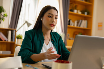 Serious asian woman working on laptop making video call in home office looking focused while talking online with business colleague on internet connection