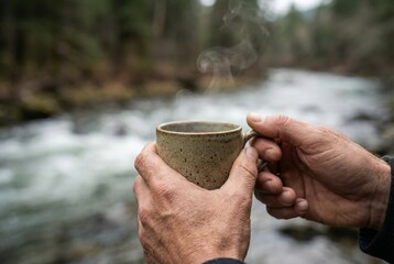 Close-up of hands holding a coffee mug with river water blurred in background