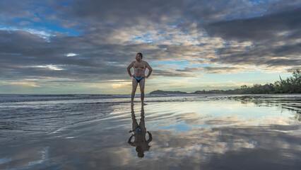 A man in swimming trunks stands on the ocean shore at dawn. Hands on his waist, looking away. The...