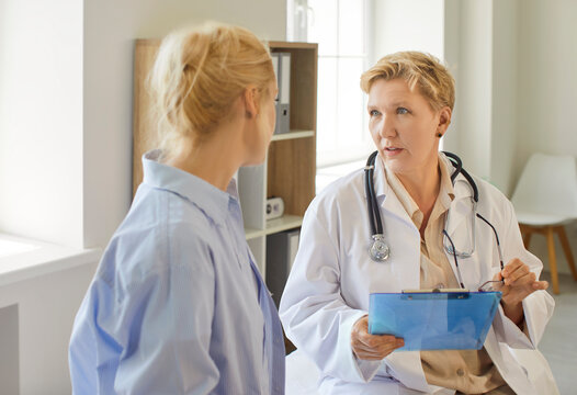 Female doctor leads a consultation with a patient in bright medical clinic, reviewing notes on a clipboard and giving professional advice. Doctor and patient engage in supportive conversation.