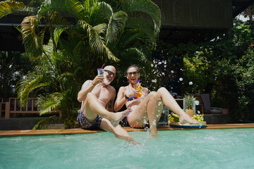 A man and a woman sit by the pool with cocktails and fruit.