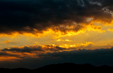 Dramatic sunset sky with golden sun rays breaking through dark clouds over mountain silhouette