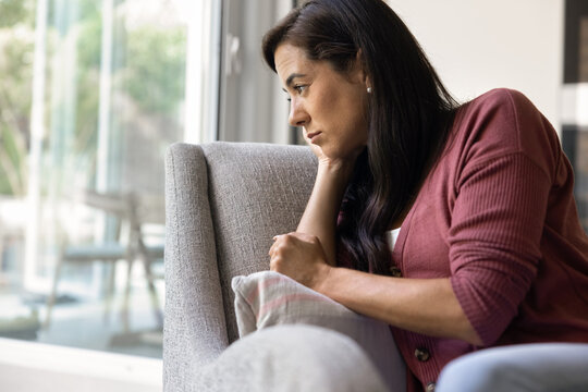 Thoughtful adult 40s woman sitting on couch near window, resting head on hand with sad expression, feeling loneliness, depression, emotional struggle in difficult life moments or situation. Break up