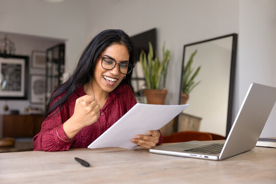 Happy excited young adult Indian girl in stylish glasses getting formal letter with good news, reading document at home work table with computer, making yes hand winner gesture