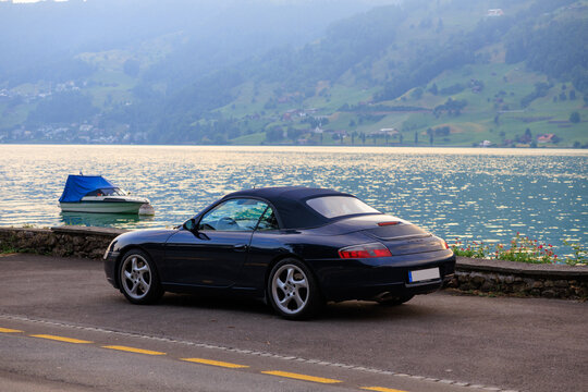 Arth, Switzerland - July 5, 2025: Blue Porsche 911 convertible next to Lake Zug (Zugersee) and boat in canton of Schwyz in Switzerland