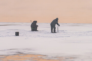 A fisherman drills a hole on the ice in winter - winter fishing. The shore of the frozen Yenisei is a Siberian river. The danger of melting ice on the river or thin ice. High quality