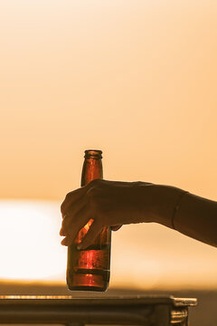 A bottle of beer at sunset at sunrise in a woman's hand. In the evening, a woman drinks beer on the beach, holding it with her hand, relaxing and relaxing on a sunny orange evening