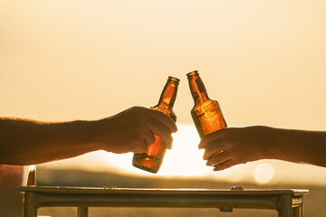 Two cold, steamed beer bottles in their hands clink glasses in close-up on the beach, against the background of the setting sun at sunset, sand and sea. Vacation and travel thirst quenching. High