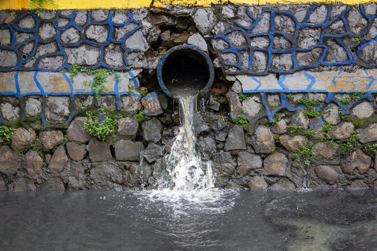 Liquid waste sewage flowing from concrete pipe on stone river wall into dark grey murky water canal with stone texture.