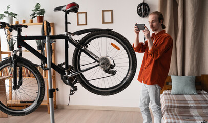 Young long-haired man in orange shirt repairing bicycle making photo for mechanic at home