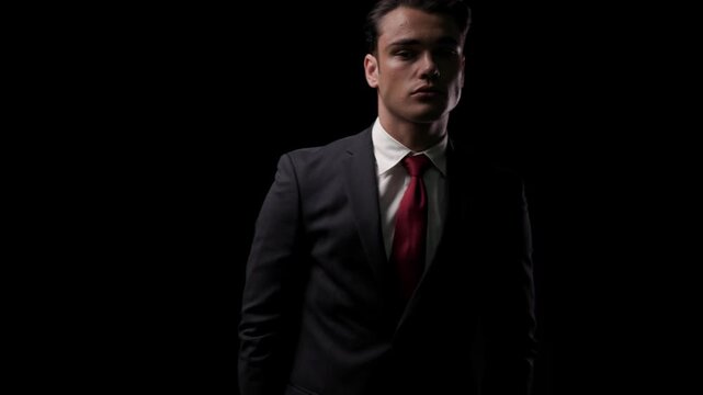 Confident man in suit standing, looking at camera, turning head to side, rotating body, and eventually facing back on black background
