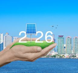 Fototapeta premium 2026 white text with solar cell on green grass field in hands over city tower, skyscraper and river with birds, Happy new year 2026 green ecology and saving energy concept