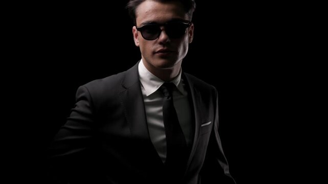 Fashion man taking off sunglasses with both hands, revealing face, looking confidently at camera and buttoning suit jacket on black background.