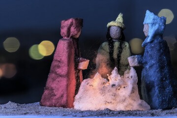 Three figures made of sand stand around a fire at night holding cups. The scene shows distant lights in a dark background